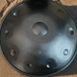 May include: A dark gray handpan instrument with a central tone field and several surrounding tone fields. The instrument has a smooth, metallic finish and is set against a sandy background. The word "FOUND" is printed on the instrument.