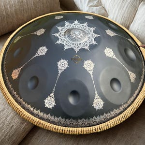 May include: A dark gray handpan drum with a decorative white mandala design in the center and other white floral patterns. The drum has a woven border and the word "FOSSE" is visible. This musical instrument is designed for melodic percussion.