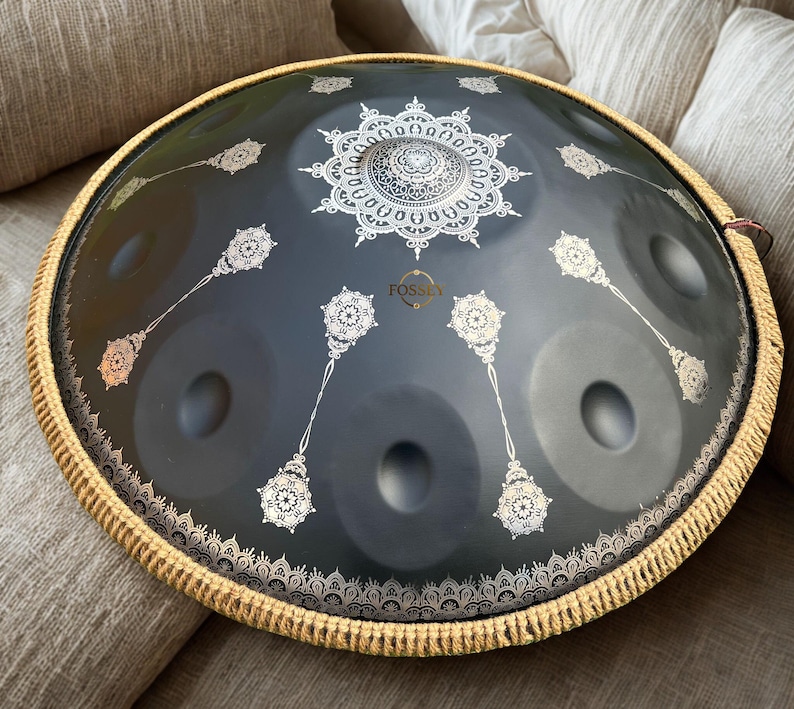 May include: A dark gray handpan drum with a decorative white mandala design in the center and other white floral patterns. The drum has a woven border and the word "FOSSE" is visible. This musical instrument is designed for melodic percussion.