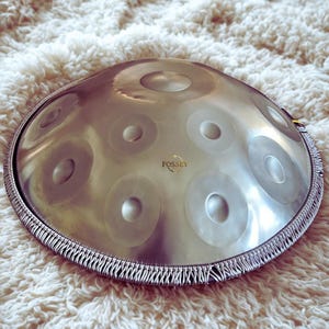May include: A silver-colored handpan instrument with multiple circular indentations on its surface. The instrument has a woven border and the word "FOSSEY" is printed on the surface. The handpan is resting on a white, fluffy surface.
