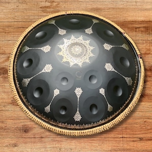 May include: A dark gray handpan with a decorative gold design. The instrument has a circular shape with multiple circular indentations and a woven border. The design includes a central mandala and floral patterns.