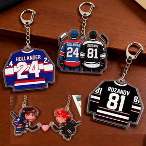 May include: Acrylic hockey jersey keychains featuring the names and numbers of players. The keychains are in blue, white, and black. One keychain shows two players holding hockey sticks. The keychains are attached to silver keyrings.