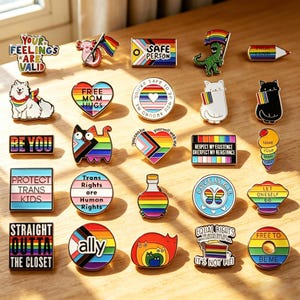 May include: A collection of colorful enamel pins with various designs and text, including rainbows, flags, cats, and phrases like "Safe Person," "Be You," and "Love is All." The pins are arranged on a wooden surface.