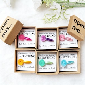 May include: A selection of belly button lint brushes presented in small cardboard boxes. Each brush features a colourful fluffy head and a matching handle. The boxes display the text "For the person who has EVERYTHING".