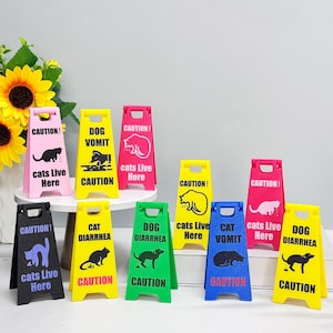 May include: A collection of colorful, miniature caution signs. Each sign features a warning about cats or dogs, with phrases like "Dog Vomit" and "Cats Live Here." The signs are in various colors, including pink, yellow, green, and blue.