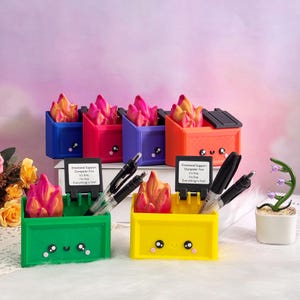 Puede incluir: Una colección de organizadores de escritorio coloridos con forma de contenedores de basura, cada uno con una escultura en forma de llama en su interior. Los organizadores son de color azul, rojo, morado, naranja, verde y amarillo. Cada contenedor tiene una cara linda.