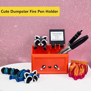 Puede incluir: Un portalápices naranja con forma de contenedor de basura con un diseño de mapache. El portalápices tiene una cara linda y contiene dos bolígrafos. El texto "Cute Dumpster Fire Pen Holder" está en la parte superior. Un letrero dice "Emotional Support Dumpster Fire. It's fine. I'm fine. Everything is fine!"