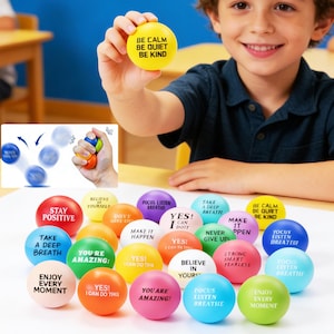 May include: A collection of colorful stress balls with positive affirmations printed on them. One yellow ball displays "BE CALM, BE QUIET, BE KIND." Other balls have phrases like "STAY POSITIVE" and "ENJOY EVERY MOMENT."