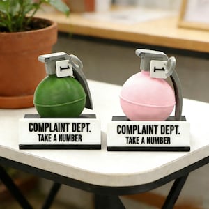 Könnte beinhalten: Zwei neuartige, granatenförmige Schreibtischaccessoires, eines grün und eines rosa, jeweils mit grauem Deckel und einem kleinen Anhänger mit der Nummer 1. Jedes steht auf einem schwarz-weißen Schild mit der Aufschrift "COMPLAINT DEPT. TAKE A NUMBER."