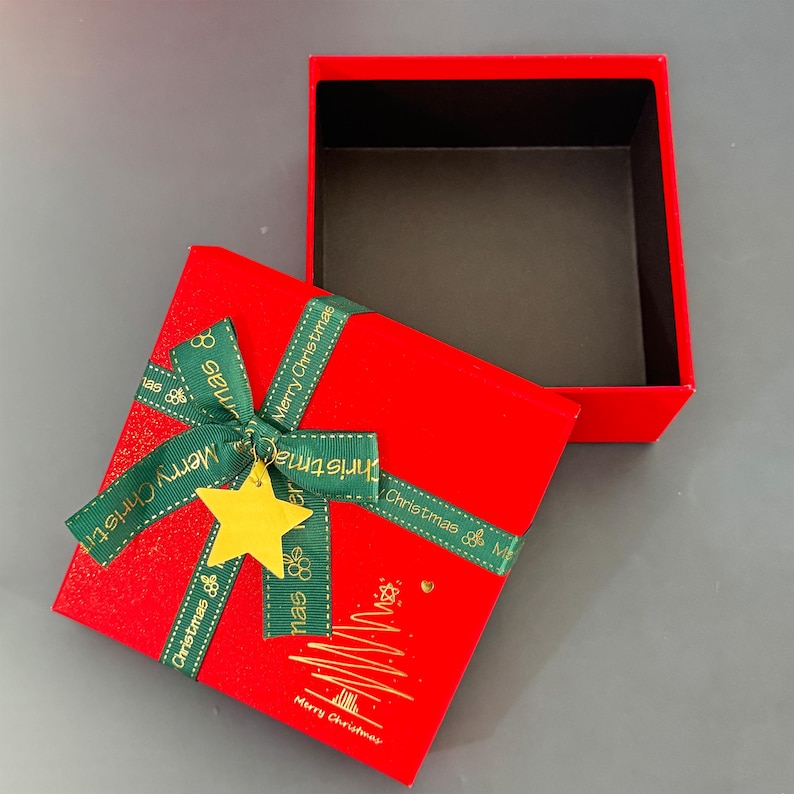 May include: A red square gift box with a matching lid, open to reveal a black interior. The box is decorated with a green ribbon that says "Merry Christmas" and a gold star charm. A gold Christmas tree is printed on the box.