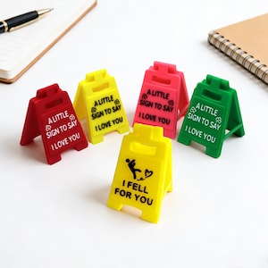 Könnte beinhalten: Eine Sammlung farbenfroher, kleiner Warnschilder. Die Schilder sind rot, gelb, rosa und grün, jedes mit einer anderen Botschaft in weißer Schrift. Ein Schild lautet "I FELL FOR YOU".
