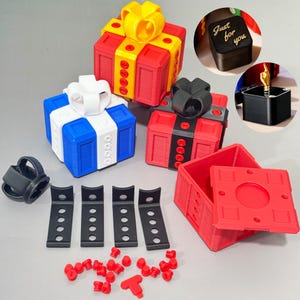 Może przedstawiać: Asortyment kolorowych, drukowanych w 3D pudełek na prezenty ze wstążkami, wraz z innymi elementami. Pudełka są czerwone, niebieskie i czarne, ze wstążkami w kolorze żółtym, białym i czarnym. W zestawie znajdują się również czarne wsporniki i czerwone łączniki. Małe czarne pudełko z napisem "Just for you".