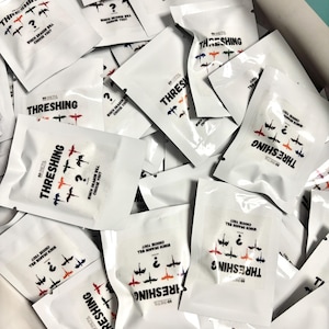 May include: A pile of white, sealed, plastic pouches with the word "THRESHING" printed on them. Each pouch features a different colourful dragon design and the text "Which dragon will choose you?"