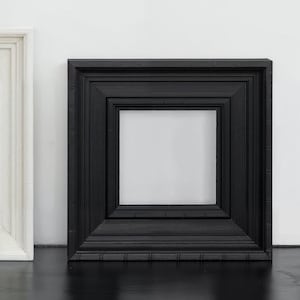 May include: A square black picture frame with a white interior. The frame has a layered design with a textured surface. A white frame is partially visible on the left. The frames are on a black surface against a white wall.