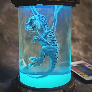 Puede incluir: Una figurita de dinosaurio azul suspendida en un frasco de vidrio transparente con una luz azul en el fondo. El dinosaurio tiene una cola larga y está enroscado en el frasco.