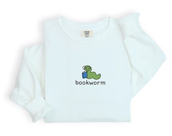 May include: White long-sleeved sweatshirt with a crew neck. The front features a cartoon bookworm wearing glasses and holding a book, with the word "bookworm" below the graphic in a black, sans-serif font.