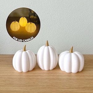 Peut inclure: Trois décorations de citrouilles blanches avec des tiges dorées sont présentées sur une surface en bois. Les citrouilles ont un design côtelé. Un encart circulaire montre trois lumières LED en forme de citrouille. Le texte dans l'encart indique "FOR LED CANDLES".