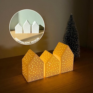 Könnte beinhalten: Drei hausförmige Kerzenhalter aus Keramik mit gepunkteten Mustern, die ein warmes Leuchten ausstrahlen. Die Kerzenhalter sind beige. Ein kleiner Weihnachtsbaum befindet sich im Hintergrund. Der Text "Minimalistic Design" befindet sich in einem Kreis.