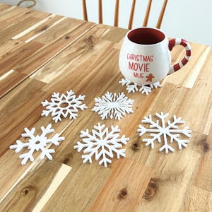 May include: Set of six white snowflake-shaped coasters with a red mug that says "Christmas Movie Mug" and a gingerbread man graphic on a wooden table.