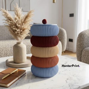 Pode incluir: Uma pilha de cinco recipientes circulares decorativos, canelados, em tons de azul, bordô, bege e ferrugem. Um pequeno objeto redondo de cor bordô está no topo. Os recipientes estão sobre uma mesa de mármore branco.