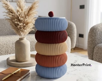May include: A stack of five decorative, ribbed, circular containers in shades of blue, burgundy, tan, and rust. A small, round, burgundy-colored object sits on top. The containers are on a white marble table.