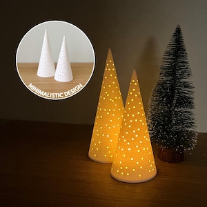 Puede incluir: Dos adornos de árbol de Navidad iluminados, en forma de cono, con un patrón de puntos. Los árboles están iluminados desde dentro, emitiendo un brillo cálido. A la derecha hay un pequeño árbol de Navidad verde oscuro. Incluye el texto "MINIMALISTIC DESIGN".
