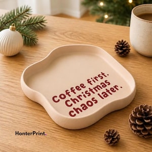 Könnte beinhalten: Ein beigefarbenes Keramiktablett mit gewelltem Rand und dem dunkelroten Text "Coffee first. Christmas chaos later.". Das Tablett hat eine glatte, matte Oberfläche und eine flache, abgerundete Schale, die zur Aufbewahrung kleiner Gegenstände dient.