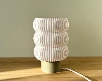 Puede incluir: Una lámpara de mesa blanca y acanalada con un diseño apilado y redondeado. La lámpara se asienta sobre una pequeña base verde claro y tiene un cable de alimentación blanco. La lámpara está sobre una superficie de madera.