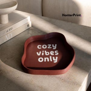 May include: A dark red, organic-shaped tray with raised edges featuring the white text "cozy vibes only" printed in the center. The tray has a textured surface and is positioned near a stack of books and a bowl, with the brand name HonterPrint visible.