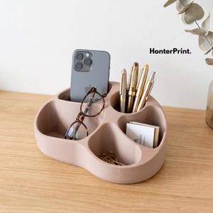 Op de afbeelding: Een matte beige bureau-organizer met vier afgeronde vakken met een grijze smartphone, een bril met schildpadpatroon, vier metalen pennen, een stapel witte visitekaartjes en diverse gouden paperclips, met de tekst HonterPrint zichtbaar.