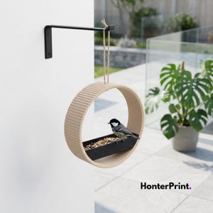 May include: A beige, circular bird feeder with a black feeding tray, suspended by twine from a black metal bracket. A small bird perches on the tray filled with seeds. The feeder is mounted on a white wall. The text "HonterPrint." is visible.