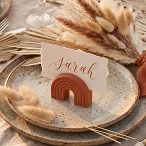 May include: A terracotta rainbow place card holder with a card inscribed with "Sarah" in cursive. The holder sits on a speckled ceramic plate, with dried flowers in the background. The scene evokes a rustic, bohemian style, ideal for a wedding or event.