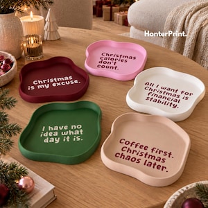 Könnte beinhalten: Fünf wellenförmige Tabletts in Pink, Dunkelrot, Weiß, Grün und Hellbeige. Jedes Tablett trägt einen aufgedruckten Text: Christmas calories don't count, Christmas is my excuse, All I want for Christmas is financial stability, I have no idea what day it is und Coffee first. Christmas chaos later.