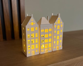 May include: Three white paper houses with cut-out windows and a light inside each house. The houses are arranged in a row on a wooden surface.