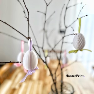 Puede incluir: Huevos de Pascua de papel blanco con cintas moradas y rosadas colgando de ramas desnudas. El texto "HonterPrint." es visible en la esquina inferior derecha de la imagen.