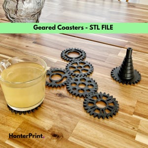 May include: A set of six black 3D printed coasters shaped like gears, with one coaster shaped like a cone. The coasters are arranged in a line on a wooden table, with a glass of tea on one of the gear-shaped coasters. The text "Geared Coasters - STL FILE" is displayed in green at the top of the image.