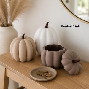 Può includere: Tre oggetti decorativi a forma di zucca e un piccolo vassoio rotondo. Una zucca è beige, una bianca e una è un contenitore color tortora con coperchio. Il testo HonterPrint è visibile. Una chiave e un anello dorati sono sul vassoio.
