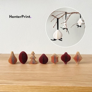 Op de afbeelding: Een verzameling papieren kerstversieringen in rood, oranje en wit. Sommige hangen aan een tak, andere staan op een houten oppervlak. De afbeelding bevat de tekst "HonterPrint."