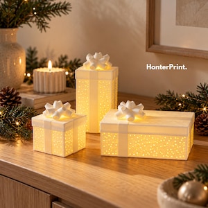 Könnte beinhalten: Drei weiße geschenkboxförmige Lampen mit einem perforierten Muster, das ein warmes gelbes Licht ausstrahlt. Jede Box ist mit einer weißen Schleife versehen. Der Text HonterPrint. ist in der oberen rechten Ecke des Bildes sichtbar.