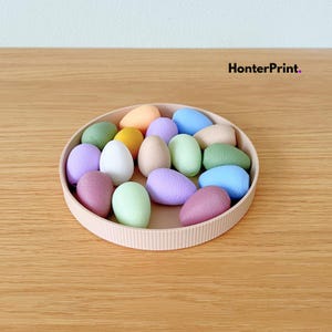 Peut inclure: Un assortiment d'objets en forme d'œufs aux couleurs pastel dans un plateau rond rose clair. Les œufs sont de différentes couleurs pastel, notamment rose, vert, bleu et jaune. Le plateau est posé sur une surface en bois, avec le texte "HonterPrint.".