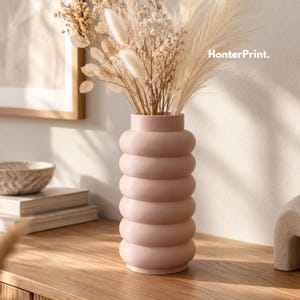 Puede incluir: Un jarrón cilíndrico de color rosa claro mate con una textura apilada de nervaduras o anillos en todo el cuerpo. El jarrón está lleno de un ramo de hierbas secas de color beige y plumas de pampa. El texto HonterPrint es visible en la esquina superior derecha.