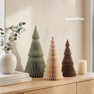 May include: Three decorative honeycomb paper trees in sage green, cream, and dark brown. The trees are arranged next to a white ceramic vase with eucalyptus branches, two stacked books, and a white pillar candle. The text HonterPrint. is visible.