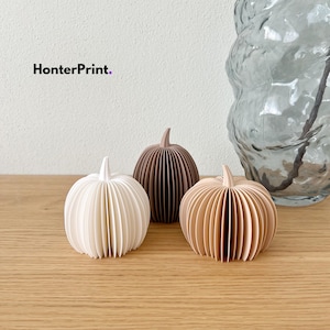 Puede incluir: Tres adornos decorativos con forma de calabaza y diseño plisado tipo acordeón. Uno es blanco, otro marrón oscuro y otro beige claro. Cada uno presenta un pequeño tallo en la parte superior. El texto HonterPrint es visible en la esquina superior izquierda.