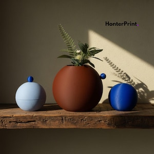 May include: Three spherical decorative objects on a wooden shelf. A light blue lidded container, a brown vase with greenery, and a blue sphere. The brown vase has a small blue handle. The text "HonterPrint" is in the upper right corner.