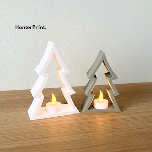 May include: Two Christmas tree-shaped candle holders, one white and one gray, each with a small, lit LED candle inside. The candle holders are made of a solid material and sit on a wooden surface. The text "HonterPrint" is in the upper left corner.