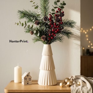 Può includere: Un vaso bianco, a gradini e plissettato, contiene un bouquet di rami di pino, foglie di eucalipto, bacche rosse e piccole pigne. Accanto al vaso si trovano una candela a colonna bianca e un piccolo ornamento a forma di casa. Il testo HonterPrint. è visibile.