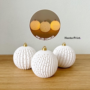 Pode incluir: Três ornamentos esféricos brancos com uma superfície nervurada texturizada e pequenos orifícios perfurados. Cada ornamento apresenta uma tampa de metal dourado no topo. Uma imagem circular inserida mostra os ornamentos iluminados para velas LED.