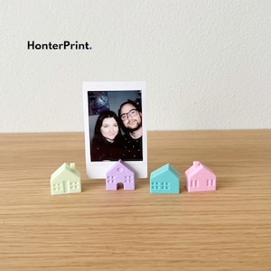 May include: A polaroid photo of a couple stands on a wooden surface, with four miniature house figurines in pastel colors. The houses are light green, lavender, teal, and pink. The text "HonterPrint." is in the upper left corner.