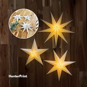 Op de afbeelding: Decoratieve stervormige lichten met een warme gloed, gemonteerd op een houten muur. De sterren hebben een witte buitenkant en een gele binnenkant, waardoor een zachte, uitnodigende sfeer ontstaat. Een kleinere inzet toont extra sterdecoraties. De tekst "HonterPrint" is inbegrepen.