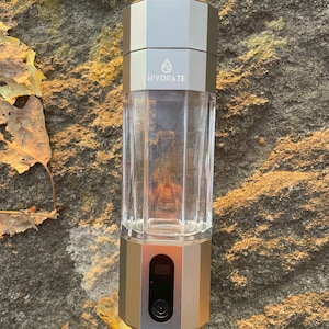 May include: A gray and silver water bottle purifier with a hexagonal design. The bottle has a clear, cylindrical reservoir and a dark gray base. The brand name 'Hydrate' is subtly printed on the bottle. The device appears to be portable and suitable for outdoor use.